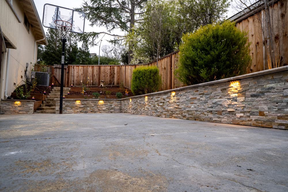 Backyard patio with stone walls, basketball hoop, and landscaped greenery at dusk.