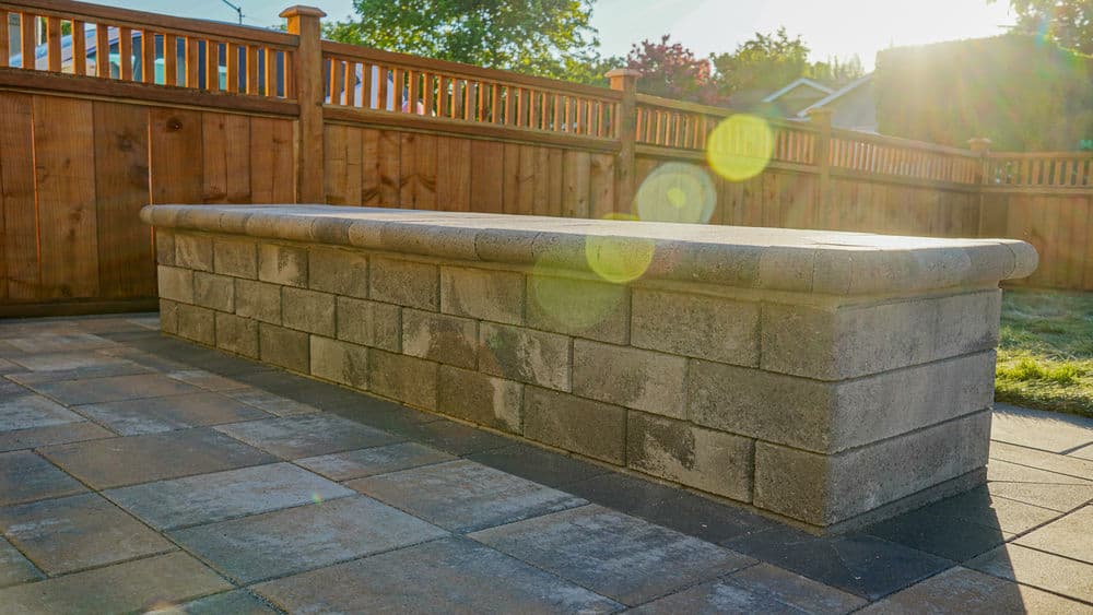 Outdoor stone bench against a wooden fence in a landscaped backyard setting.